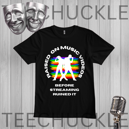 A black graphic t-shirt displayed on a gray wood-plank background, featuring a black circular text design that reads 'RAISED ON MUSIC VIDEOS BEFORE STREAMING RUINED IT' around a black silhouette of two people, with a rainbow stripe pattern behind them, along with two silver comedy masks and a vintage microphone graphic.