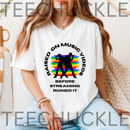 A woman wearing a white graphic T-shirt with a black circular design featuring two dancing figures over a rainbow and the text 'RAISED ON MUSIC VIDEOS BEFORE STREAMING RUINED IT.' The photo is taken in a softly lit indoor setting with a blanket and neutral wall in the background.