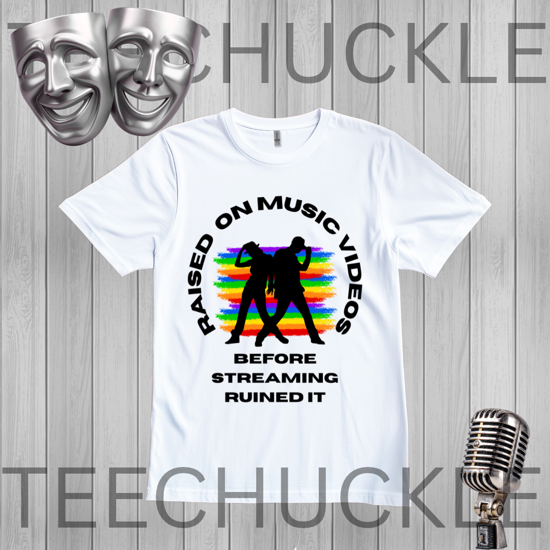 A white graphic t-shirt displayed on a gray wood-plank background, featuring a black circular text design that reads 'RAISED ON MUSIC VIDEOS BEFORE STREAMING RUINED IT' around a black silhouette of two people, with a rainbow stripe pattern behind them, along with two silver comedy masks and a vintage microphone graphic.