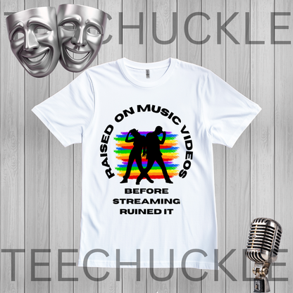 A white graphic t-shirt displayed on a gray wood-plank background, featuring a black circular text design that reads 'RAISED ON MUSIC VIDEOS BEFORE STREAMING RUINED IT' around a black silhouette of two people, with a rainbow stripe pattern behind them, along with two silver comedy masks and a vintage microphone graphic.