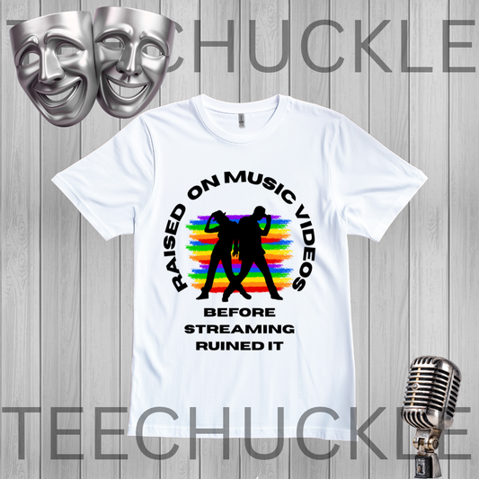 A white graphic t-shirt displayed on a gray wood-plank background, featuring a black circular text design that reads 'RAISED ON MUSIC VIDEOS BEFORE STREAMING RUINED IT' around a black silhouette of two people, with a rainbow stripe pattern behind them, along with two silver comedy masks and a vintage microphone graphic.