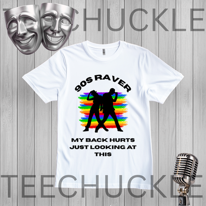 A white graphic t-shirt displayed on a gray wood-plank background, featuring a black silhouette of two people dancing in front of a rainbow stripe pattern, with the text '90S RAVER MY BACK HURTS JUST LOOKING AT THIS'. The image includes decorative silver theater masks in the top-left and a vintage silver microphone in the bottom-right