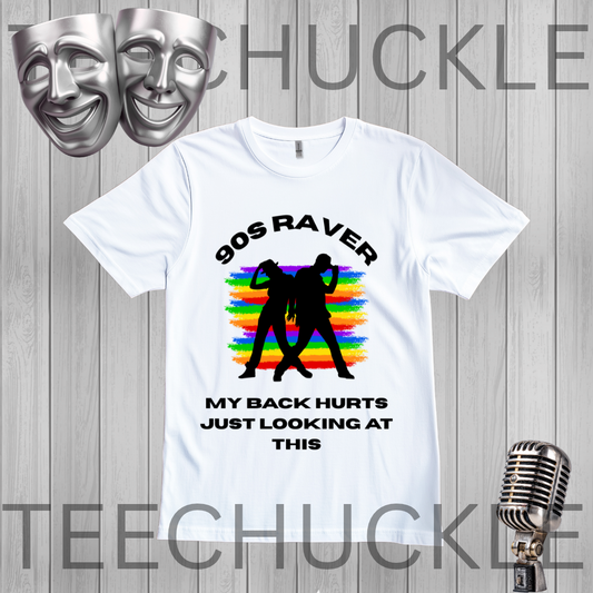 A white graphic t-shirt displayed on a gray wood-plank background, featuring a black silhouette of two people dancing in front of a rainbow stripe pattern, with the text '90S RAVER MY BACK HURTS JUST LOOKING AT THIS'. The image includes decorative silver theater masks in the top-left and a vintage silver microphone in the bottom-right