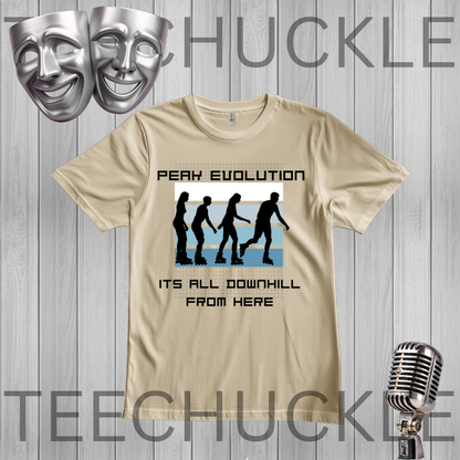 A graphic t-shirt displayed on a gray wood-plank background, featuring a 'Peaky Evolution' design with black silhouettes of figures walking downhill and the text 'ITS ALL DOWNHILL FROM HERE'. Silver theater masks and a vintage microphone are shown as decorative props