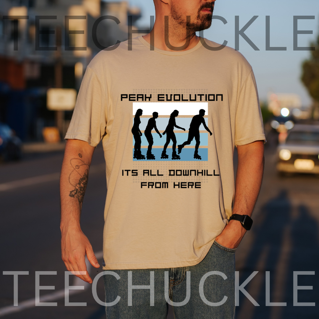 A man wearing a sand graphic t-shirt with a 'PEAK EVOLUTION' downhill silhouette design, standing outdoors on a street at sunset. 