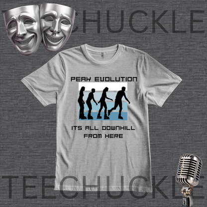 A gray graphic t-shirt displayed on a dark gray textured background, featuring a 'Peak Evolution' downhill silhouette graphic. The image includes decorative props like silver theater masks in the top-left and a vintage microphone in the bottom-right