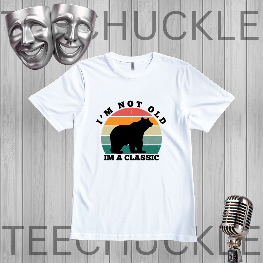 A white short-sleeve graphic t-shirt displayed on a gray wood-plank background. The shirt features a retro-style graphic with a black bear silhouette and the text 'I'M NOT OLD' above and 'I'M A CLASSIC' below, set against a striped sunset in orange, yellow, and green.
