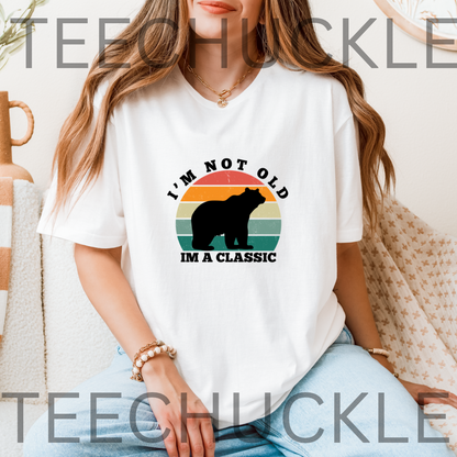 A woman wearing a white graphic t-shirt with a bear silhouette and the text 'I'M NOT OLD IM A CLASSIC' over a retro sunset design. 