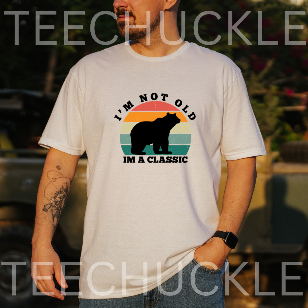 A man wearing a white graphic t-shirt with a retro-style bear design and the text 'I'M NOT OLD I'M A CLASSIC'. The background is a blurred outdoor setting, 