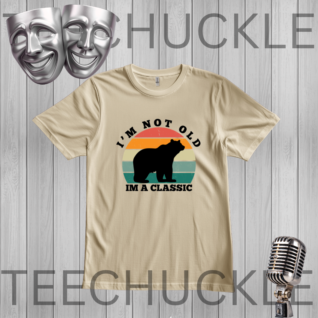 A sand graphic t-shirt displayed flat on a gray wood plank background. The shirt features a retro-style graphic with a black bear silhouette over a sunset gradient (orange, yellow, green, blue) and the text 'I'M NOT OLD IM A CLASSIC'.