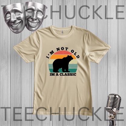 A sand graphic t-shirt displayed flat on a gray wood plank background. The shirt features a retro-style graphic with a black bear silhouette over a sunset gradient (orange, yellow, green, blue) and the text 'I'M NOT OLD IM A CLASSIC'.
