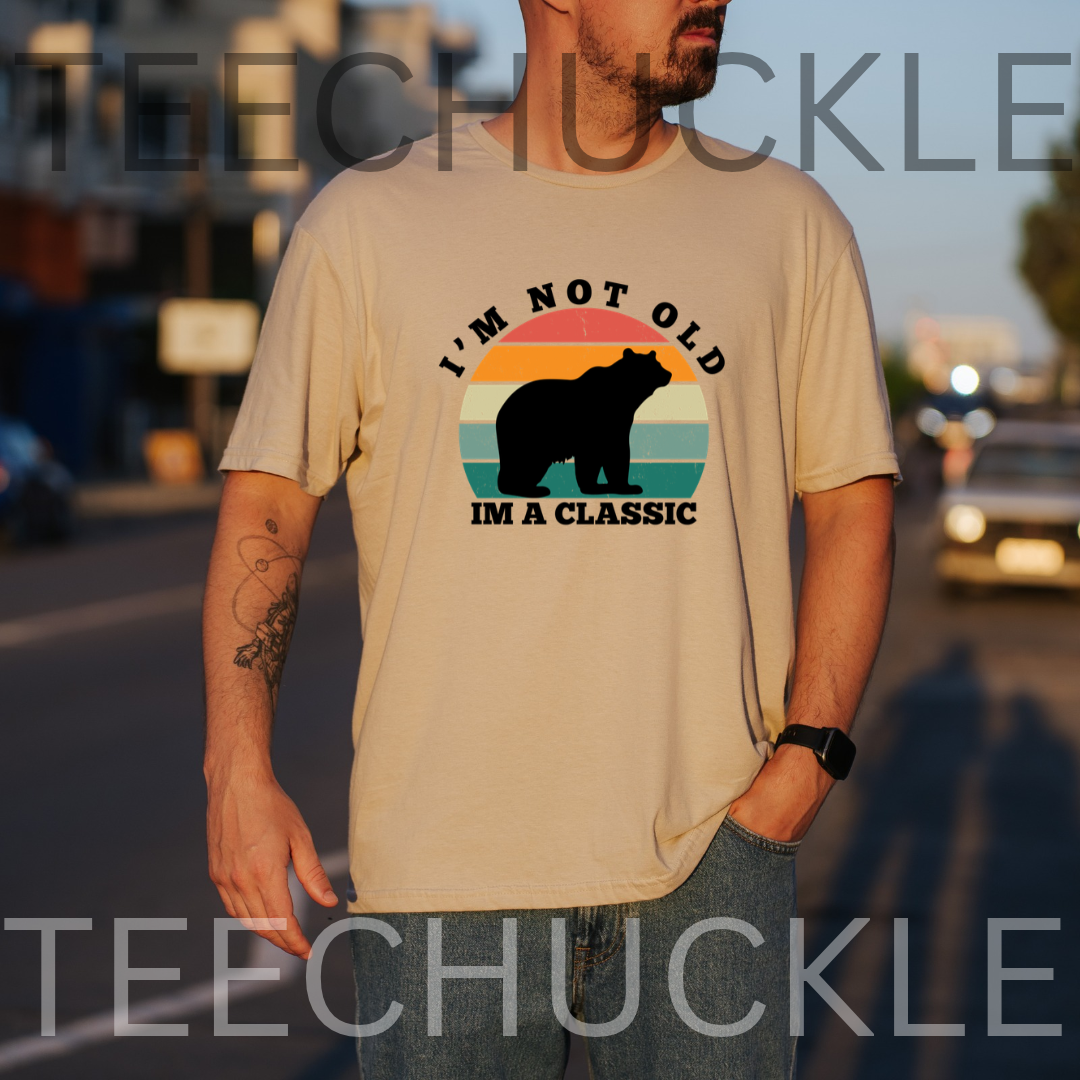 A man wearing a sand graphic t-shirt with a retro sunset bear design and the text 'I'M NOT OLD IM VINTAGE'. The background is a blurred street at sunset, and there is a large 'TEECHUCKLE' watermark overlay.