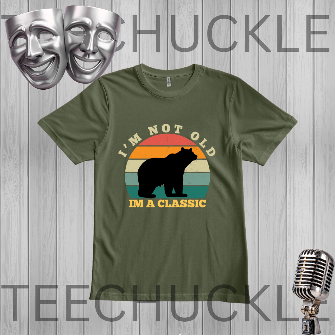 A green graphic t-shirt featuring a retro sunset design with a black bear silhouette and the text 'I'M NOT OLD IM A CLASSIC'. The t-shirt is displayed on a gray wood-plank background with decorative props including silver theater masks and a vintage microphone
