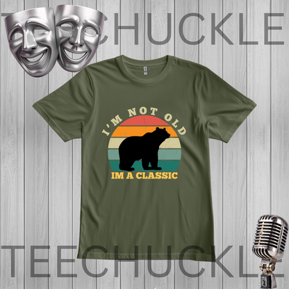 A green graphic t-shirt featuring a retro sunset design with a black bear silhouette and the text 'I'M NOT OLD IM A CLASSIC'. The t-shirt is displayed on a gray wood-plank background with decorative props including silver theater masks and a vintage microphone