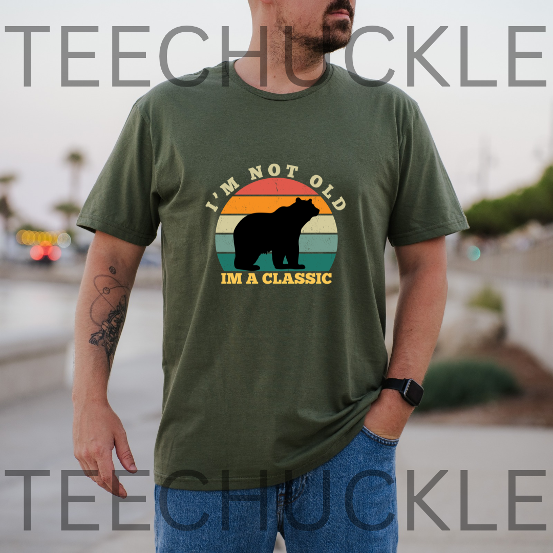 A man wearing a green graphic t-shirt with a retro sunset bear design and the text 'I'M NOT OLD I'M A CLASSIC'.and is set in an outdoor environment.