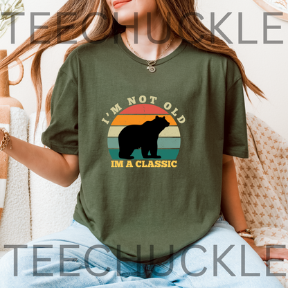 A woman wearing a green graphic t-shirt with a bear silhouette and the text 'I'M NOT OLD I'M A CLASSIC' over a retro sunset background.