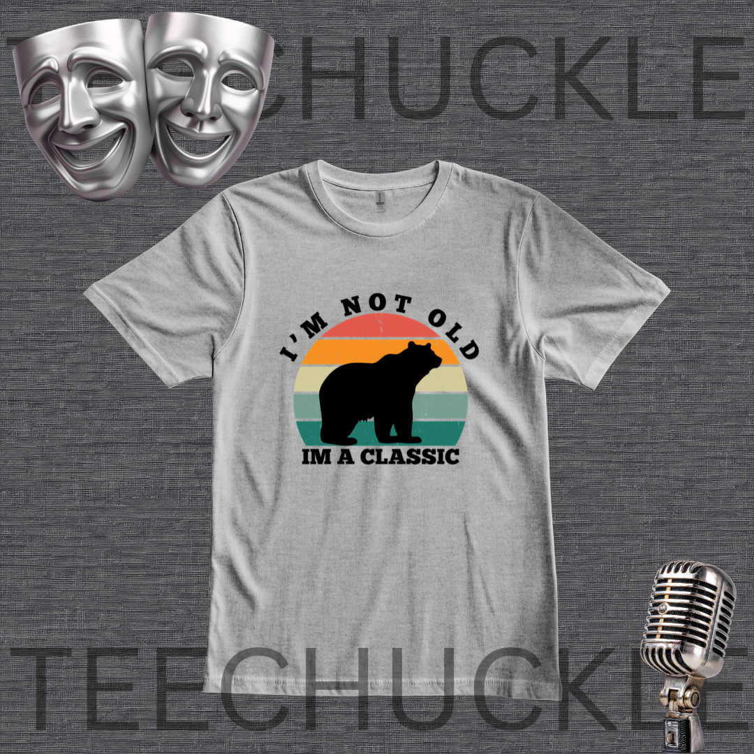 A gray graphic t-shirt featuring a retro-style bear design with the text 'I'M NOT OLD IM A CLASSIC'. The t-shirt is displayed on a dark gray textured background with decorative props including two silver theater masks, a vintage microphone