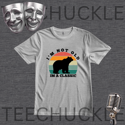 A gray graphic t-shirt featuring a retro-style bear design with the text 'I'M NOT OLD IM A CLASSIC'. The t-shirt is displayed on a dark gray textured background with decorative props including two silver theater masks, a vintage microphone