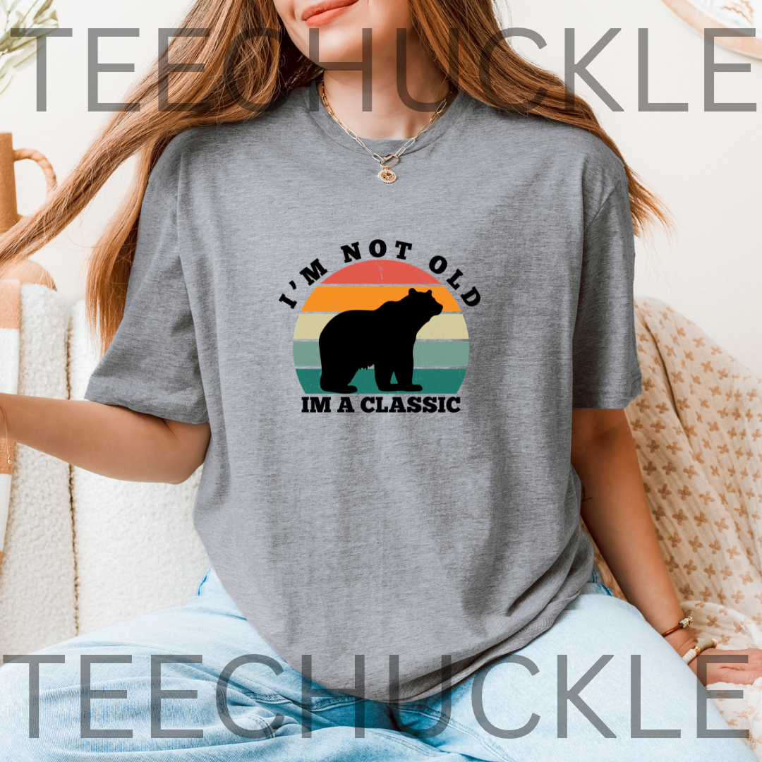A woman wearing a gray graphic t-shirt with a retro sunset bear design and the text 'I'M NOT OLD IM A CLASSIC'.