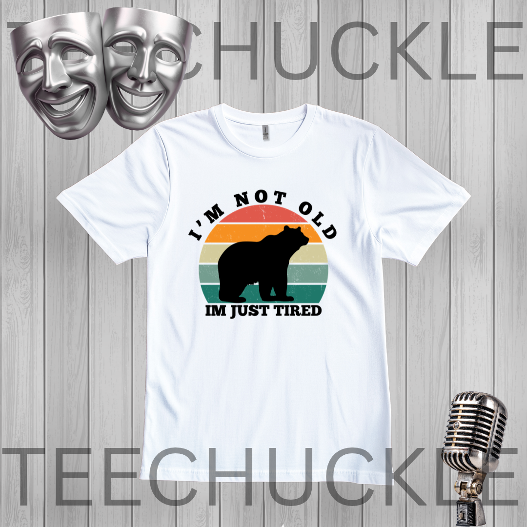 A white short-sleeve graphic T-shirt laid flat on a light gray wood-plank background. The shirt has a retro-style multicolor sunset graphic with a black bear silhouette and the text 'I'M NOT OLD IM JUST TIRED.