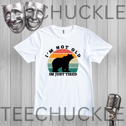 A white short-sleeve graphic T-shirt laid flat on a light gray wood-plank background. The shirt has a retro-style multicolor sunset graphic with a black bear silhouette and the text 'I'M NOT OLD IM JUST TIRED.