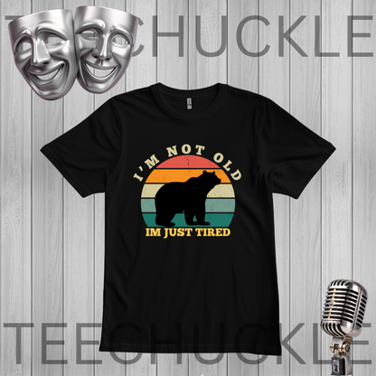 A black short-sleeve graphic t-shirt displayed on a gray wood-plank background. The shirt features a retro-style multicolor sunset graphic with a black bear silhouette and the text 'I'M NOT OLD IM JUST TIRED'.