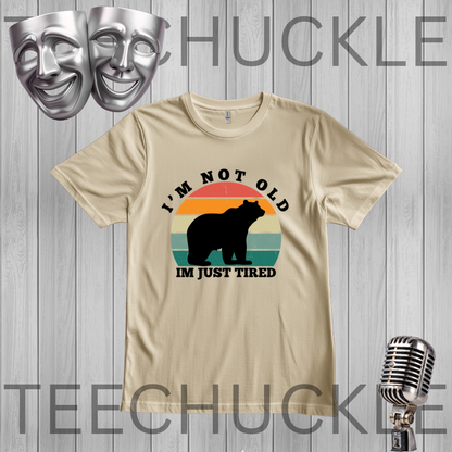 A sand graphic t-shirt featuring a black bear silhouette and a retro sunset design with the text 'I'M NOT OLD IM JUST TIRED'. The t-shirt is displayed on a gray wood-plank background with decorative props including two silver theater masks, a vintage microphone