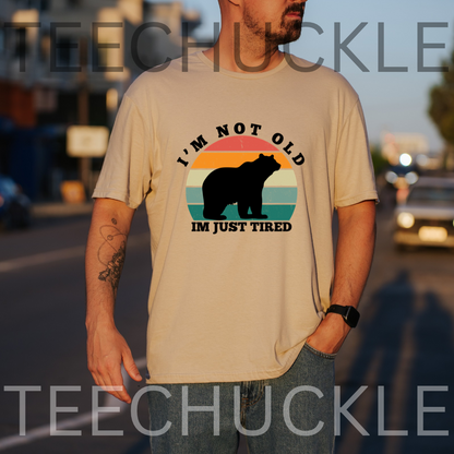 A man wearing a sand graphic t-shirt with a bear silhouette and the text 'I'M NOT OLD IM JUST TIRED' on a blurred street background.