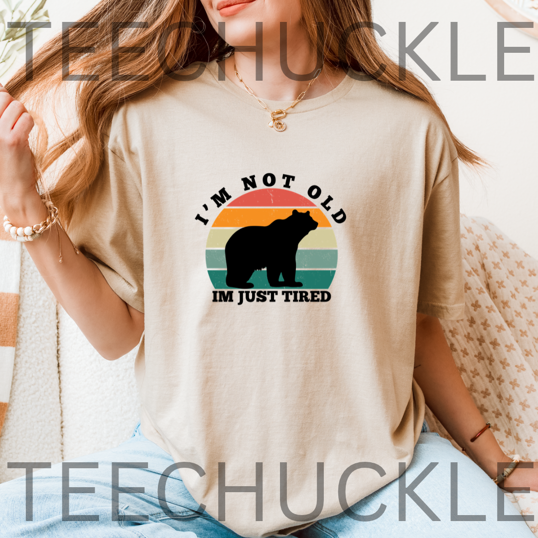 A woman wearing a sand graphic t-shirt with a retro sunset and bear design that reads 'I'M NOT OLD IM JUST TIRED'. 