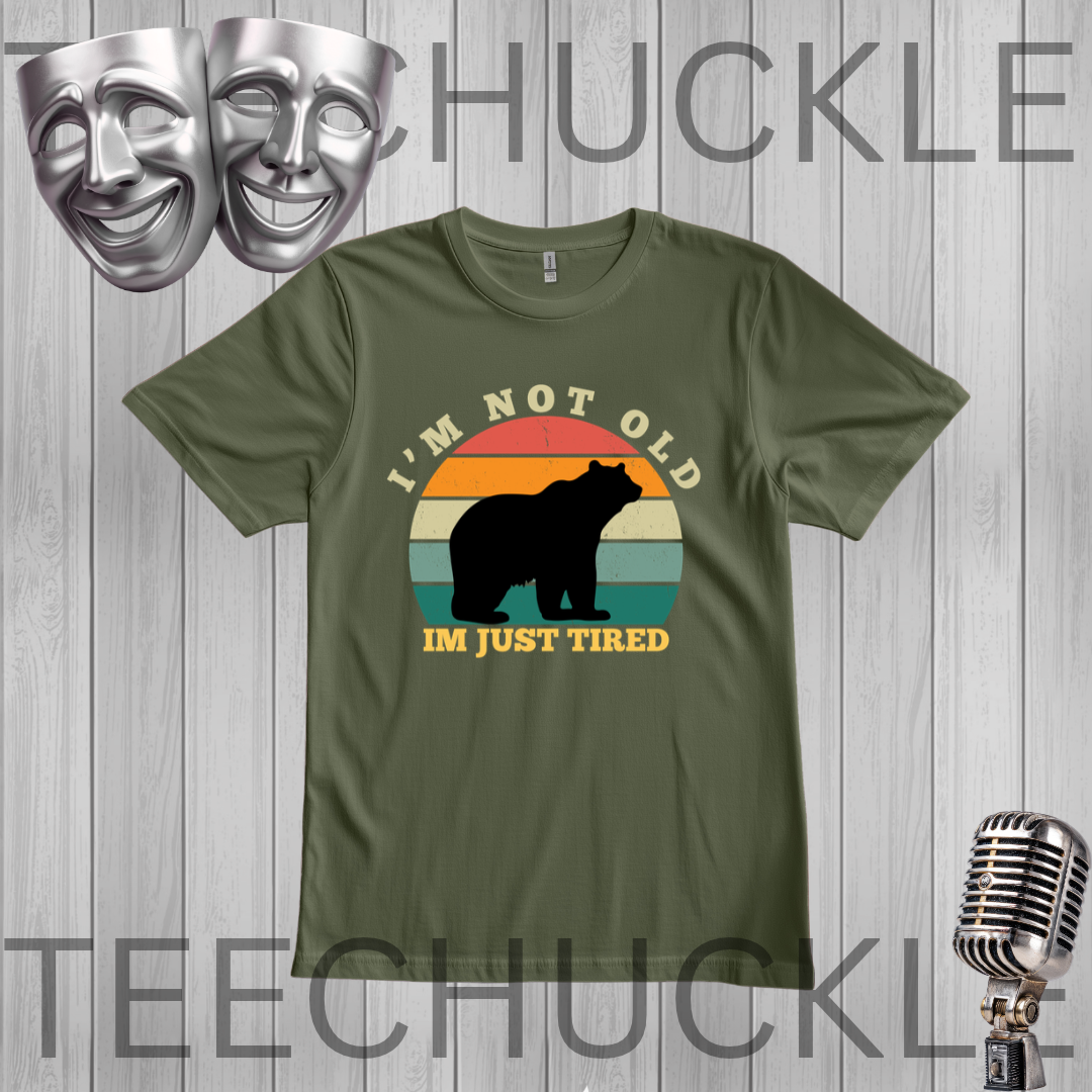 A green short-sleeve t-shirt with a retro-style graphic of a bear silhouette in front of a sunset gradient and the text 'I'M NOT OLD I'M JUST TIRED'. The t-shirt is displayed on a gray wood-plank background with vintage-style silver microphones and theater masks