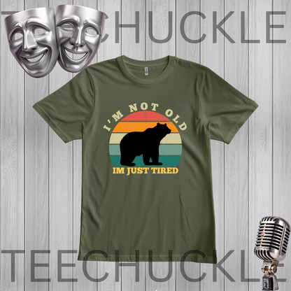 A green short-sleeve t-shirt with a retro-style graphic of a bear silhouette in front of a sunset gradient and the text 'I'M NOT OLD I'M JUST TIRED'. The t-shirt is displayed on a gray wood-plank background with vintage-style silver microphones and theater masks