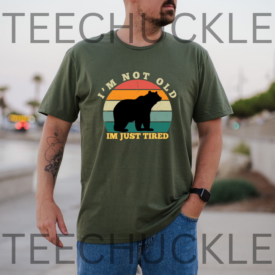 A man wearing an olive green graphic t-shirt with a bear silhouette and the text 'I'M NOT OLD IM JUST TIRED' over a retro sunset design. T