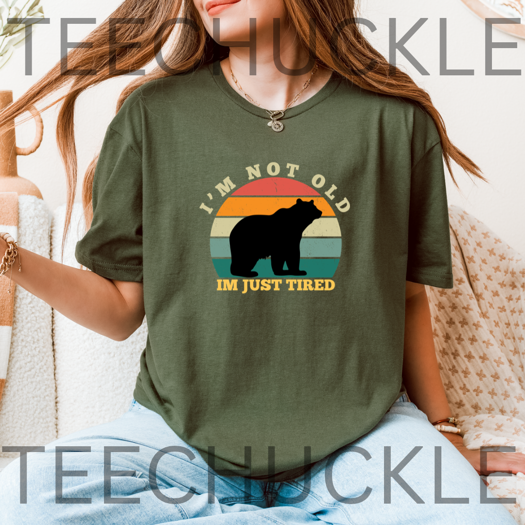A woman wearing an olive green graphic t-shirt with a bear silhouette and the text 'I'M NOT OLD IM JUST TIRED' on a retro sunset background.