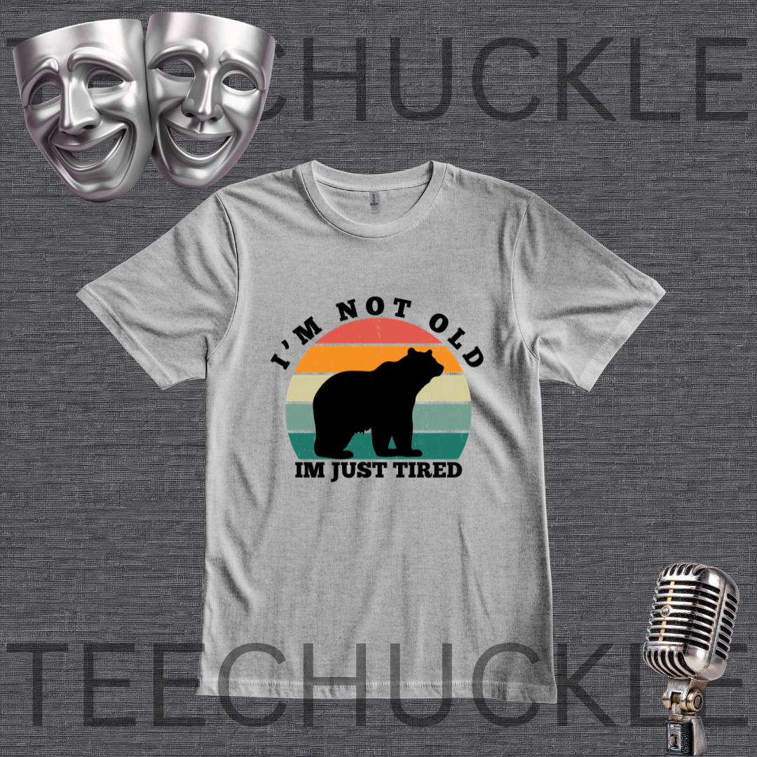 A gray graphic t-shirt featuring a retro-style bear silhouette and the text 'I'M NOT OLD IM JUST TIRED' on a dark gray textured background. The image includes decorative props like silver theater masks and a vintage microphone