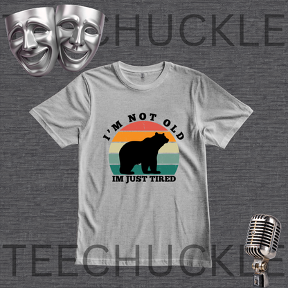 A gray graphic t-shirt featuring a retro-style bear silhouette and the text 'I'M NOT OLD IM JUST TIRED' on a dark gray textured background. The image includes decorative props like silver theater masks and a vintage microphone