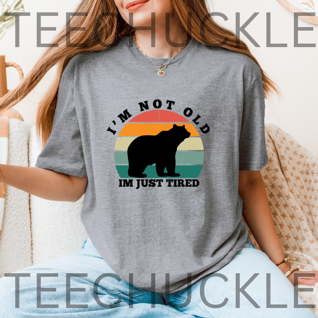A woman wearing a gray graphic t-shirt with a retro sunset bear design and the text 'I'M NOT OLD IM JUST TIRED'. 