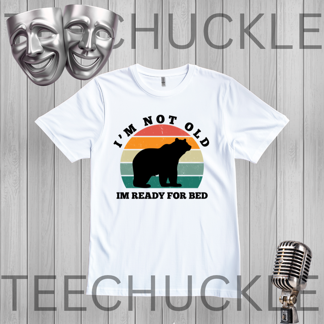 A white short-sleeve graphic T-shirt laid flat on a gray wood-plank background. The shirt has a retro sunset-style circular graphic with a bear silhouette and the text 'I'M NOT OLD IM READY FOR BED.