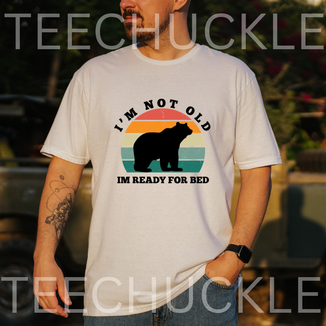 A man wearing a white graphic t-shirt with a retro-style bear design and the text 'I'M NOT OLD IM READY FOR BED'. The background is a blurred outdoor setting, 