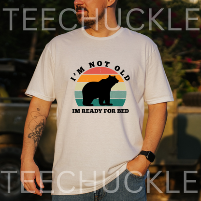 A man wearing a white graphic t-shirt with a retro-style bear design and the text 'I'M NOT OLD IM READY FOR BED'. The background is a blurred outdoor setting, 