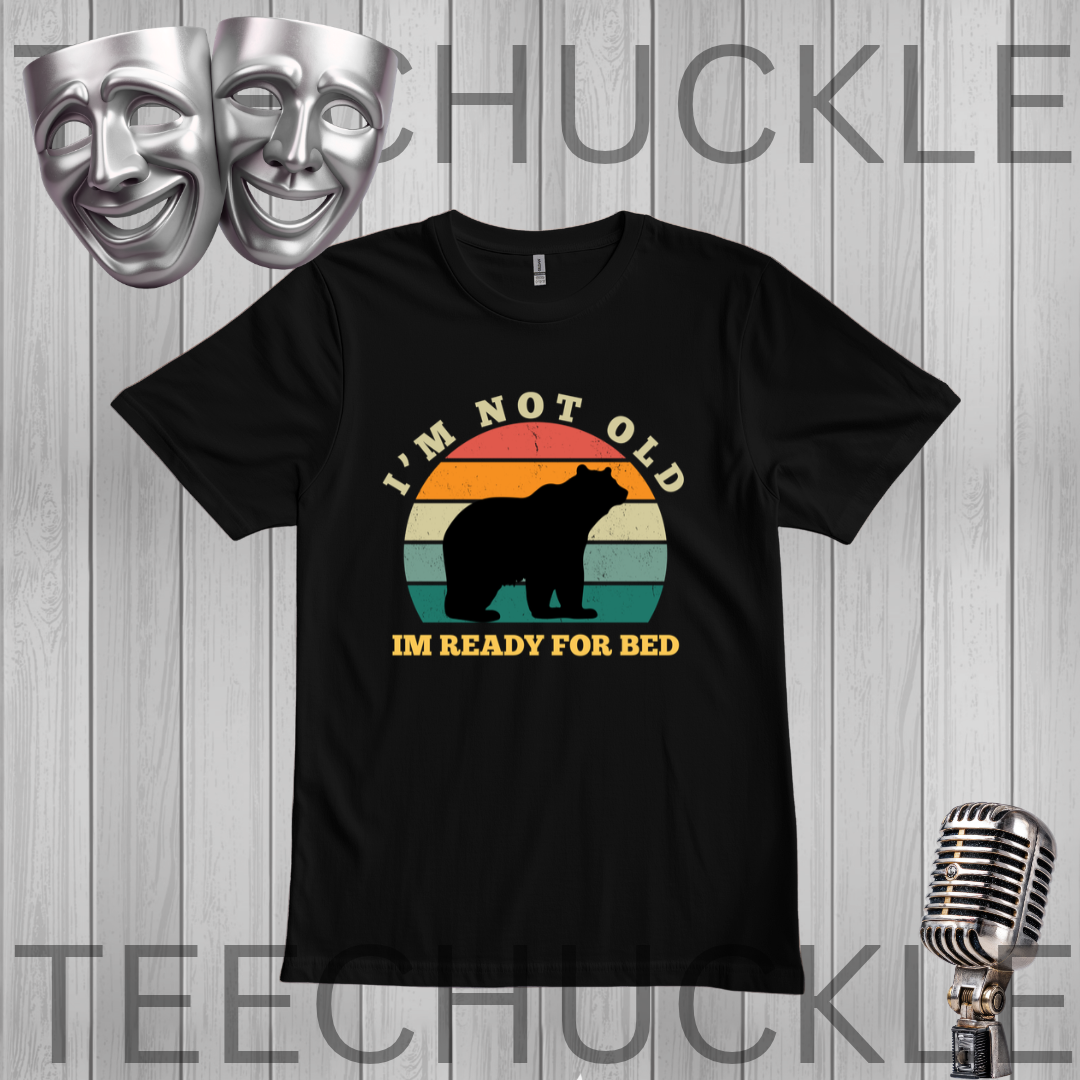 A black short-sleeve graphic t-shirt displayed on a gray wood plank background. The shirt features a retro-style sunset graphic with a bear silhouette and the text 'I'M NOT OLD IM READY FOR BED'.