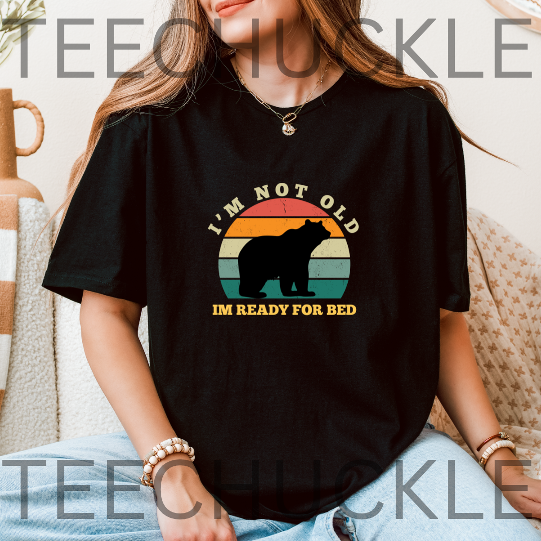 A woman wearing a black graphic t-shirt with a retro sunset bear design and the text 'I'M NOT OLD I'M READY FOR BED'. 