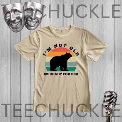 A sand graphic t-shirt featuring a bear silhouette and a retro sunset design with the text 'I'M NOT OLD IM READY FOR BED'. The t-shirt is displayed on a gray wood-plank background with decorative props including two silver theater masks, a vintage-style microphone,
