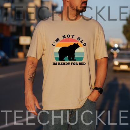 A man wearing a beige graphic t-shirt with a bear silhouette and the text 'I'M NOT OLD IM READY FOR BED' on a blurred street background. 