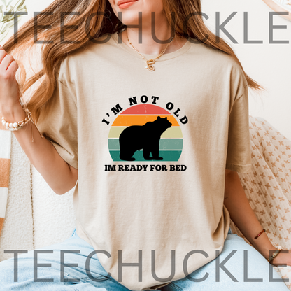 A woman wearing a beige graphic t-shirt with a retro sunset bear design and the text 'I'M NOT OLD IM READY FOR BED'. 