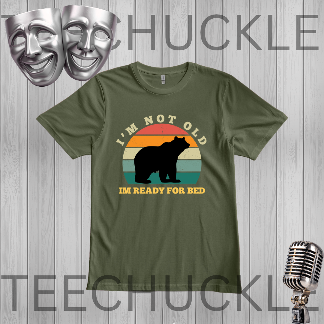 A green short-sleeve t-shirt with a retro-style graphic of a bear silhouette in front of a sunset, accompanied by the text 'I'M NOT OLD' and 'I'M READY FOR BED'. The t-shirt is displayed on a gray wood-plank background with decorative props including two silver comedy masks at the top left and a vintage-style microphone at the bottom right.