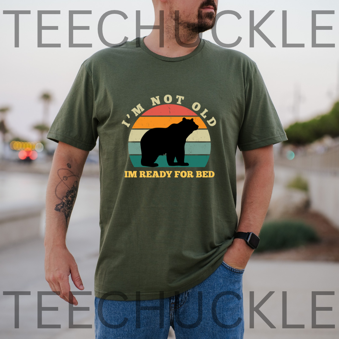 A man wearing a green graphic t-shirt with a bear silhouette and the text 'I'M NOT OLD IM JUST TIRED' over a retro sunset design.