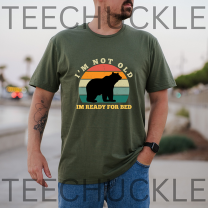 A man wearing a green graphic t-shirt with a bear silhouette and the text 'I'M NOT OLD IM JUST TIRED' over a retro sunset design.