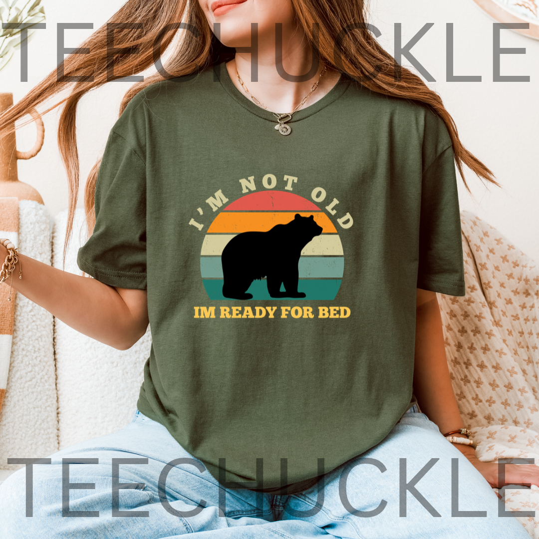 A woman wearing a green graphic t-shirt with a bear silhouette and the text 'I'M NOT OLD IM READY FOR BED' over a retro sunset background. 