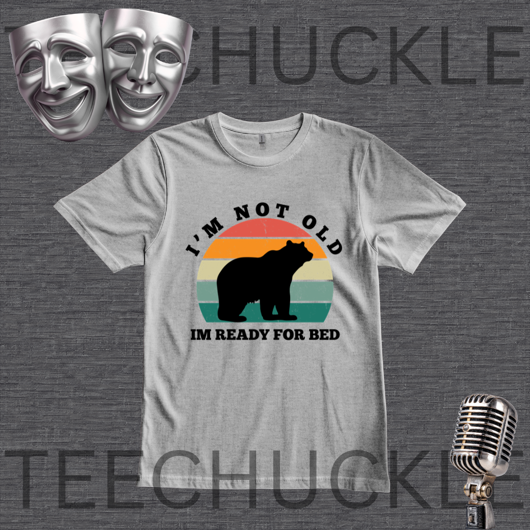 A  gray graphic t-shirt featuring a retro-style bear silhouette design with the text 'I'M NOT OLD IM READY FOR BED' on a dark gray textured background. The image includes decorative props like silver theater masks and a vintage microphone