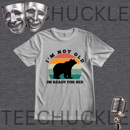 A  gray graphic t-shirt featuring a retro-style bear silhouette design with the text 'I'M NOT OLD IM READY FOR BED' on a dark gray textured background. The image includes decorative props like silver theater masks and a vintage microphone